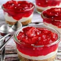 Glass jars with strawberry pretzel dessert next to spoons on a wood surface