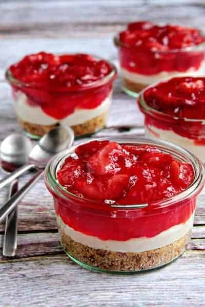 Glass jars with strawberry pretzel dessert next to spoons on a wood surface