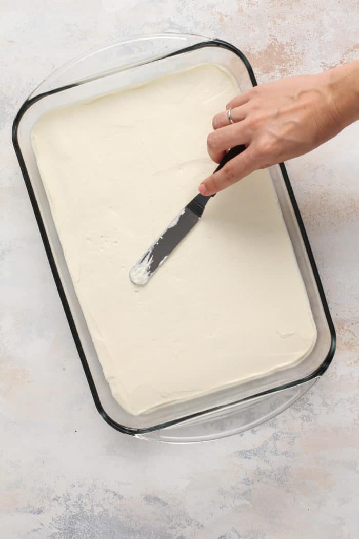 Cream cheese layer being spread over the pretzel crust in a glass baking dish.