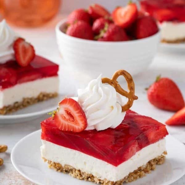 White plate with a slice of strawberry pretzel salad on it. The dessert is topped with whipped cream and a pretzel.