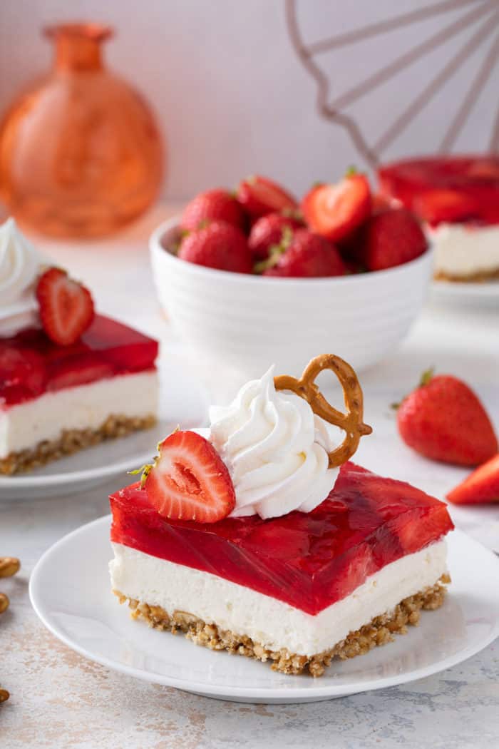 White plate with a slice of strawberry pretzel salad on it. The dessert is topped with whipped cream and a pretzel.