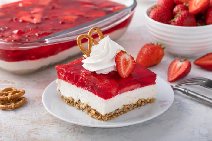 Close up of a slice of strawberry pretzel salad, topped with whipped cream and a pretzel, on a white plate.