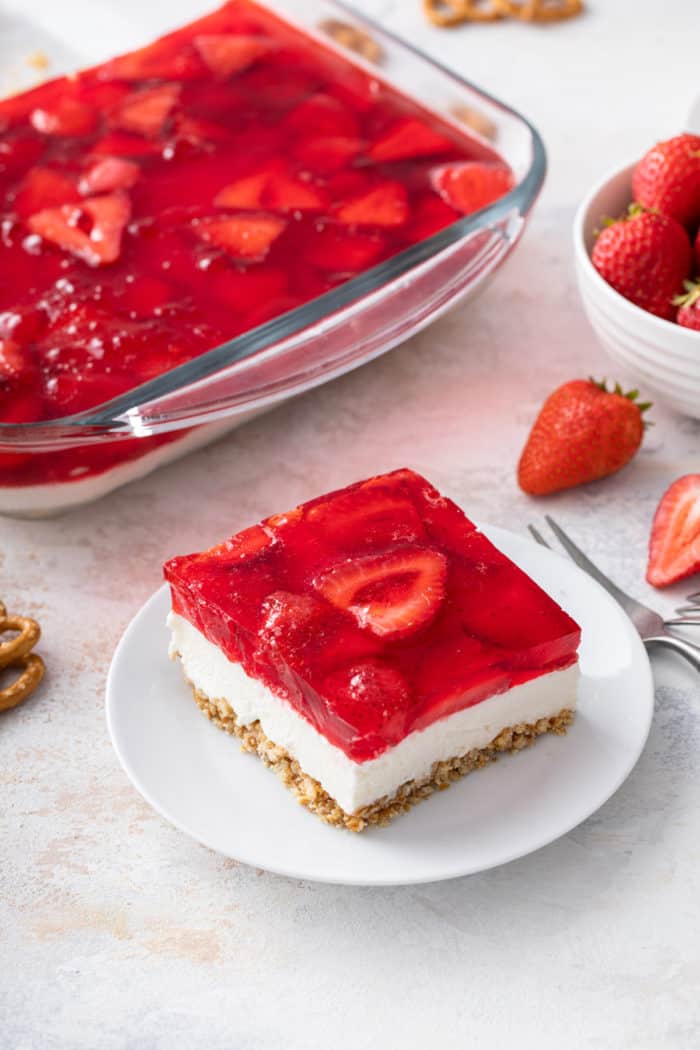 Slice of strawberry pretzel salad on a white plate. The rest of the dessert is in the background.