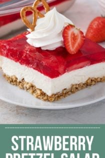 Close up of a slice of strawberry pretzel salad, topped with whipped cream and a pretzel, on a white plate. text overlay includes recipe name.