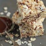 Coconut Rice Krispies stacked next to a spoon and dish of chocolate on a slate surface