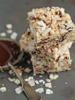 Coconut Rice Krispies stacked next to a spoon and dish of chocolate on a slate surface