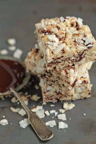 Coconut Rice Krispies stacked next to a spoon and dish of chocolate on a slate surface