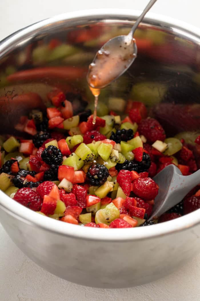 Spoon drizzling dressing over diced fresh fruit in a metal bowl.