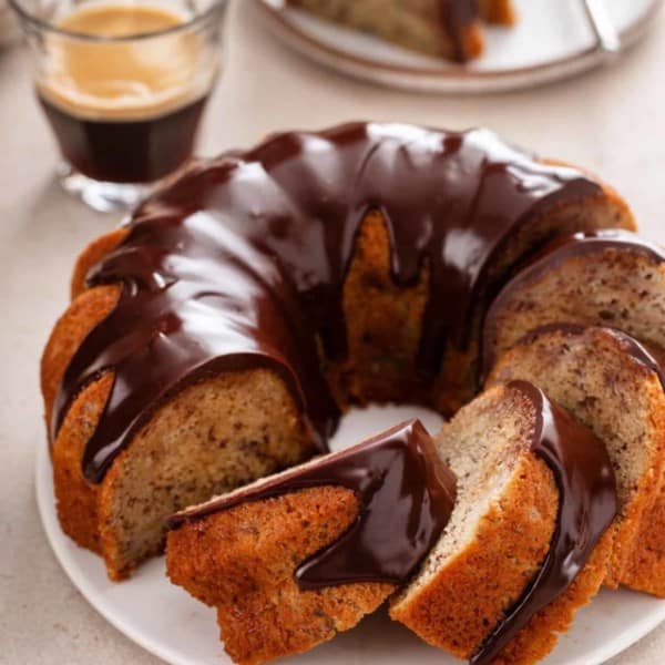 Sliced banana bundt cake topped with chocolate ganache on a white cake plate.