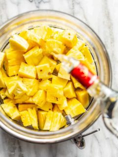 Vodka being poured over diced pineapple in a glass container