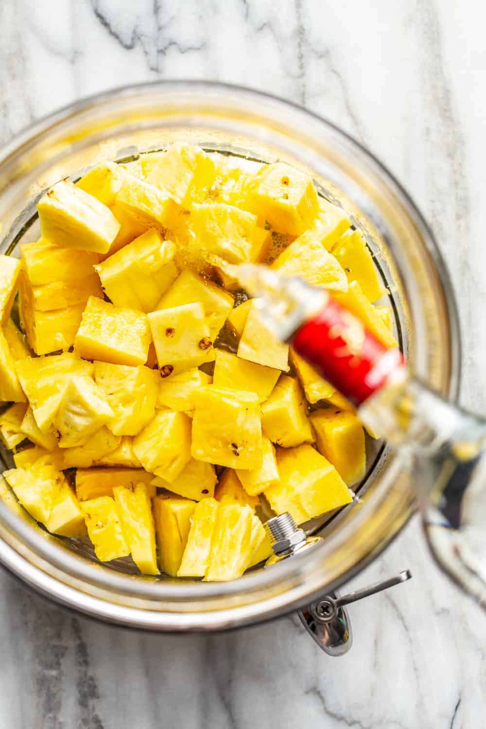 Vodka being poured over diced pineapple in a glass container