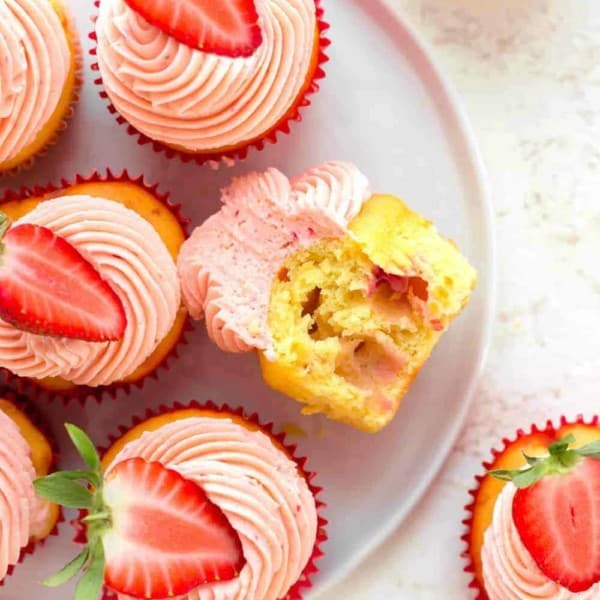 Overhead view of easy strawberry cupcake with a bite taken out of it on a white plate, surrounded by other strawberry cupcakes