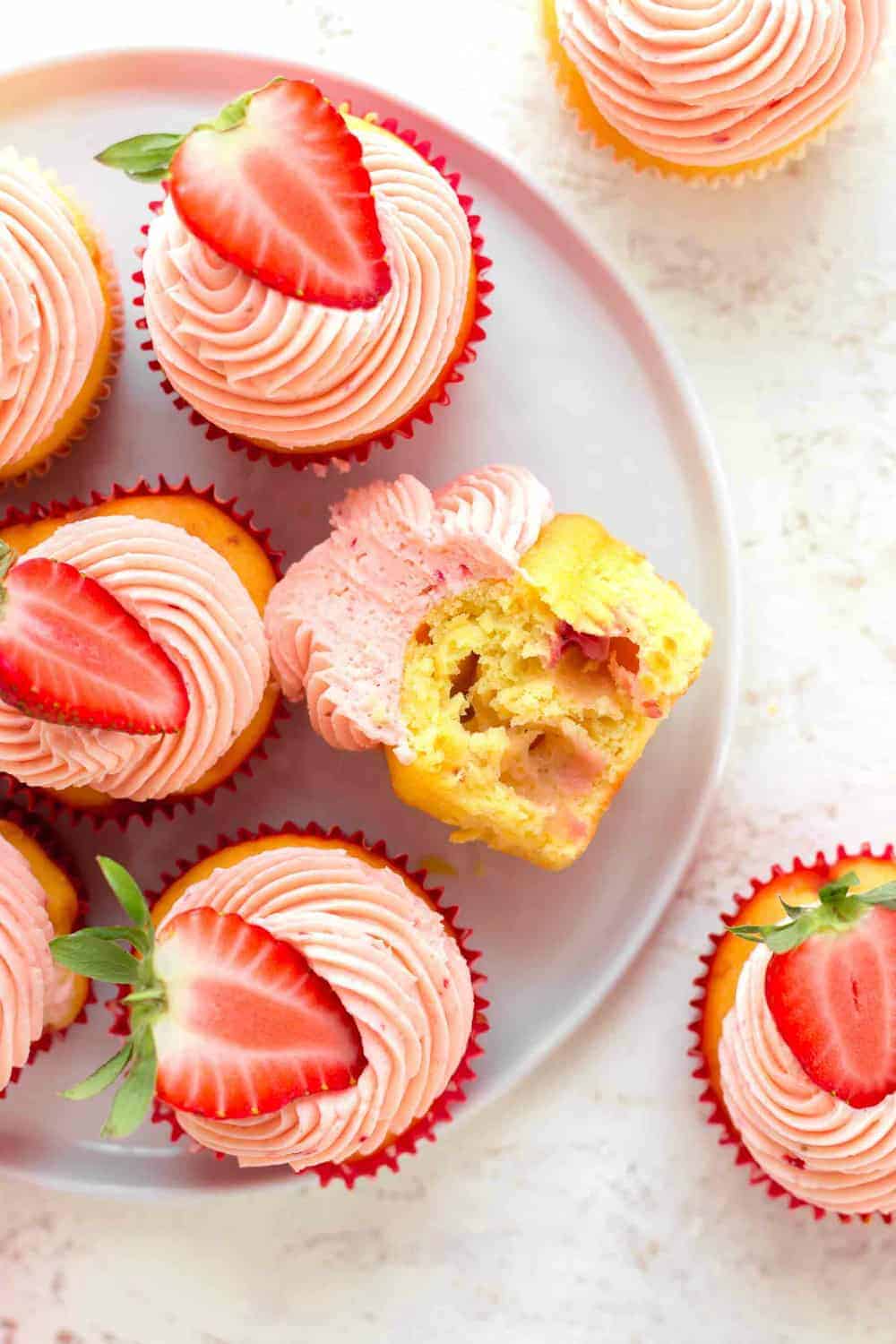 Overhead view of easy strawberry cupcake with a bite taken out of it on a white plate, surrounded by other strawberry cupcakes