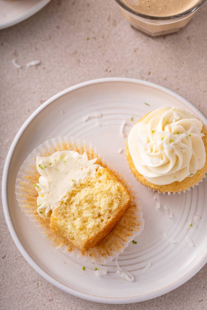 Two coconut cupcakes on a white plate. One of the cupcakes is unwrapped and cut in half.