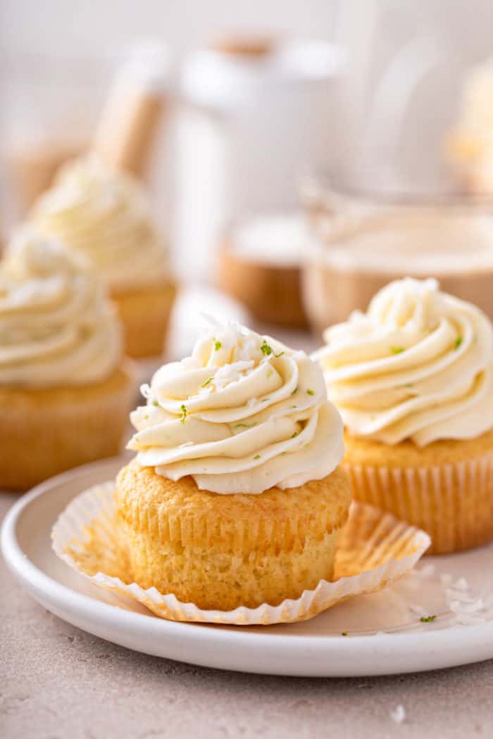 Unwrapped coconut cupcake on a white plate. More cupcakes are visible in the background.