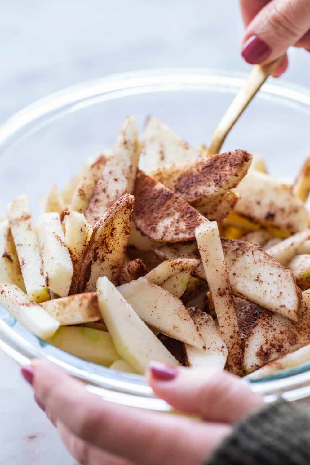 Spoon stirring sliced apples with spice blend in a glass mixing bowl
