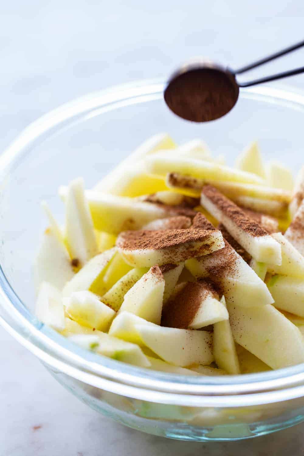 Measuring spoon sprinkling apple pie spice over sliced apples in a bowl