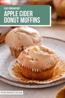 Two apple cider donut muffins on a plate. The muffin in front is unwrapped. Text overlay includes recipe name.