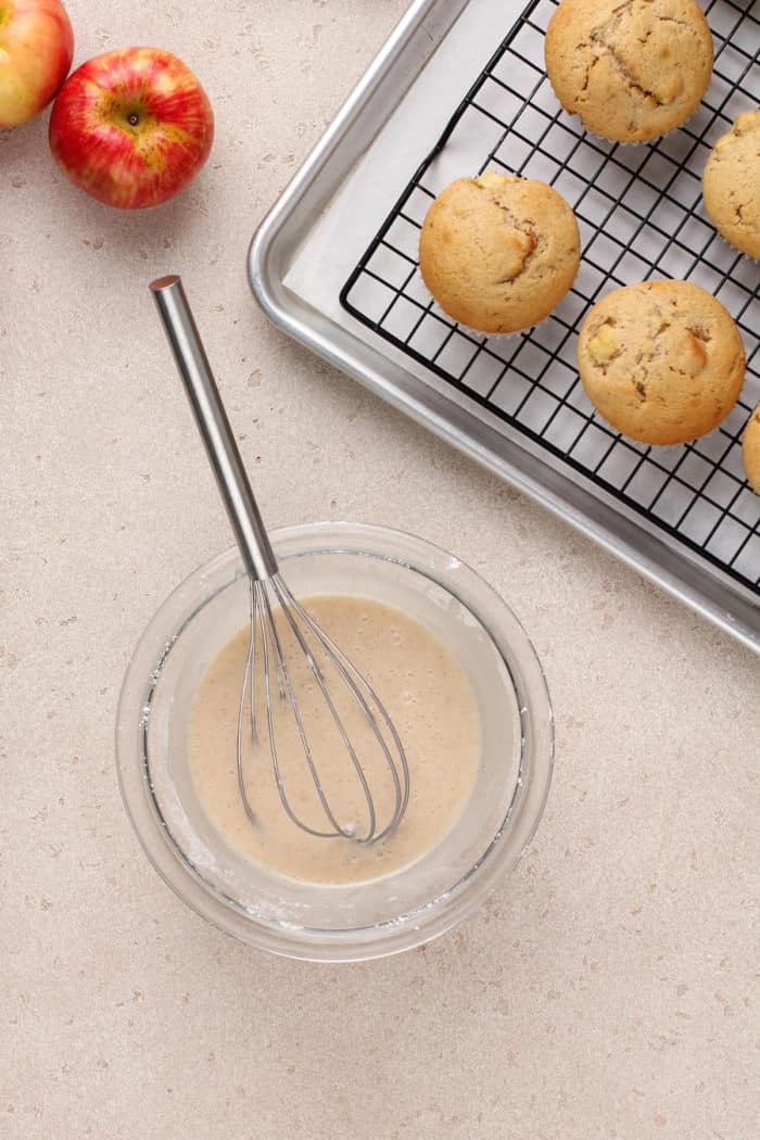 Bowl of apple cider glaze next to cooling apple cider donut muffins.