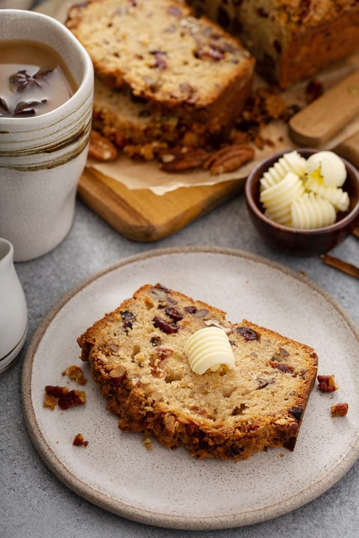 Slice of apple pie bread on a cream plate with coffee and more slices of bread on a cutting board in the background