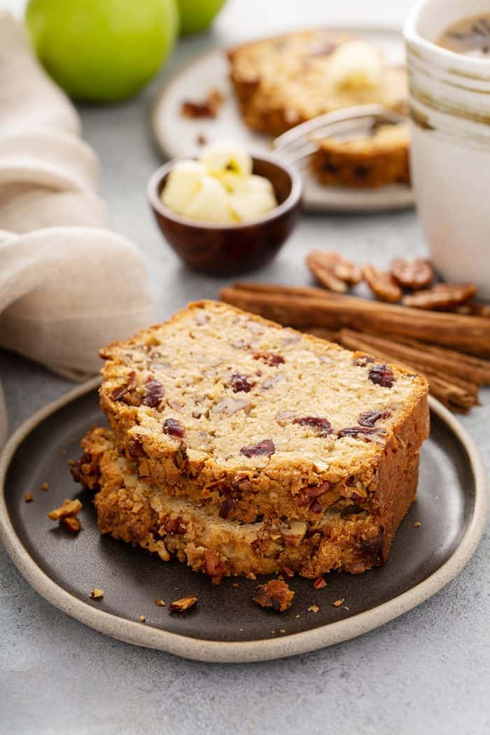 Two slices of apple pie bread stacked on a dark gray plate with cinnamon sticks and butter in the background