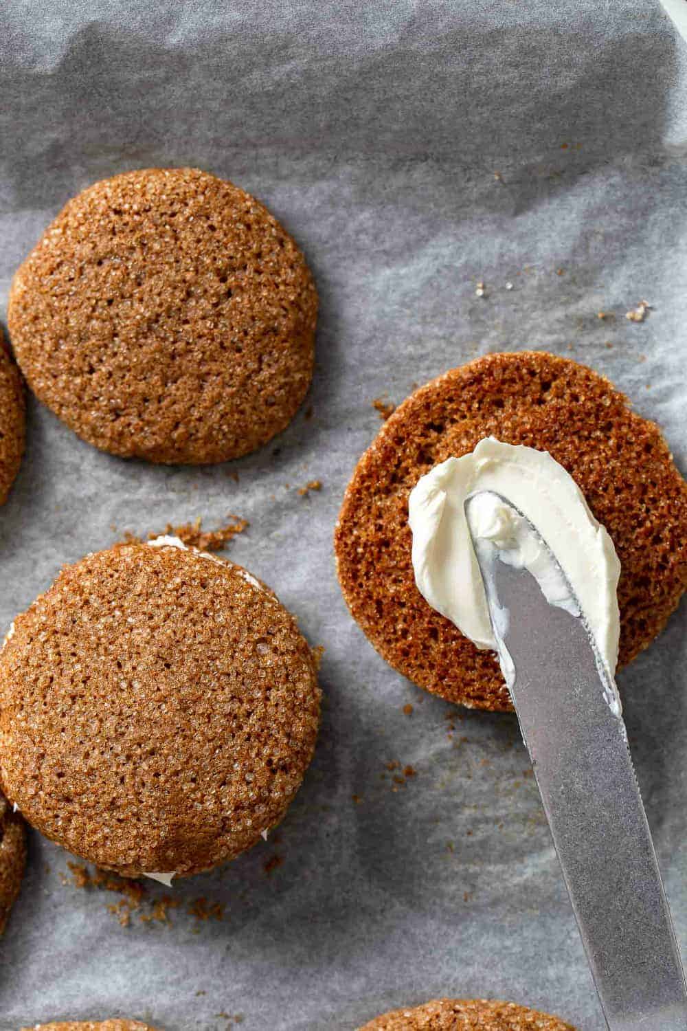 Knife spreading cream cheese filling on the bottom of a pumpkin molasses cookie