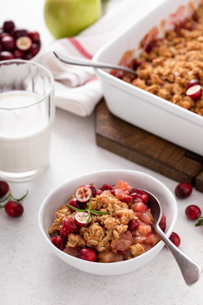 Cranberry apple crisp in a white bowl. A glass of milk and the serving dish of crisp are in the background.