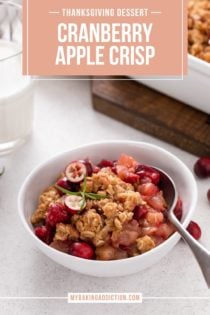Cranberry apple crisp in a white bowl. A glass of milk and the serving dish of crisp are in the background. Text overlay includes recipe name.