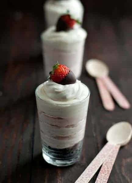 Shot glasses filled with no bake strawberry oreo cheesecake topped with a chocolate covered strawberry next to spoons