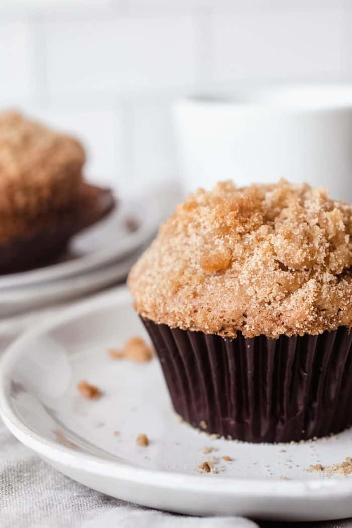 Banana crumb muffin on a white plate with additional muffins and coffee cups in the background