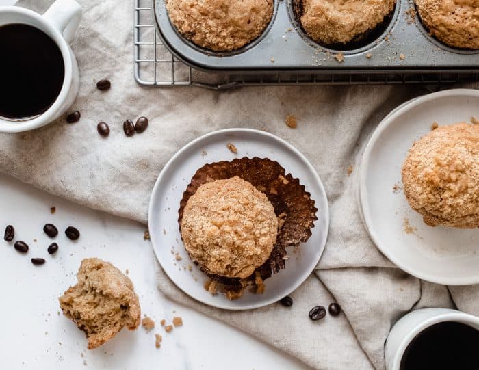 Overhead view of a banana crumb muffin on a white plate, surrounded by additional muffins, coffee and a muffin tin