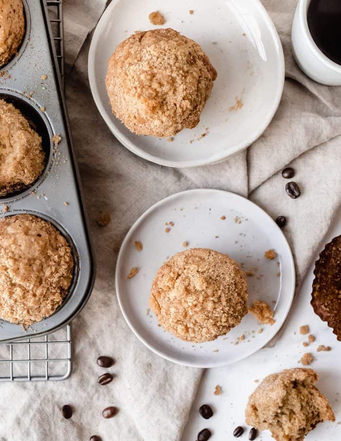 Two white plates with banana crumb muffins set on a dish towel, next to a muffin tin with more muffins