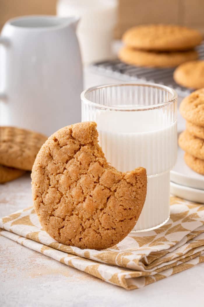 Bite taken from a peanut butter cookie that is leaning against a glass of milk, set on a napkin.