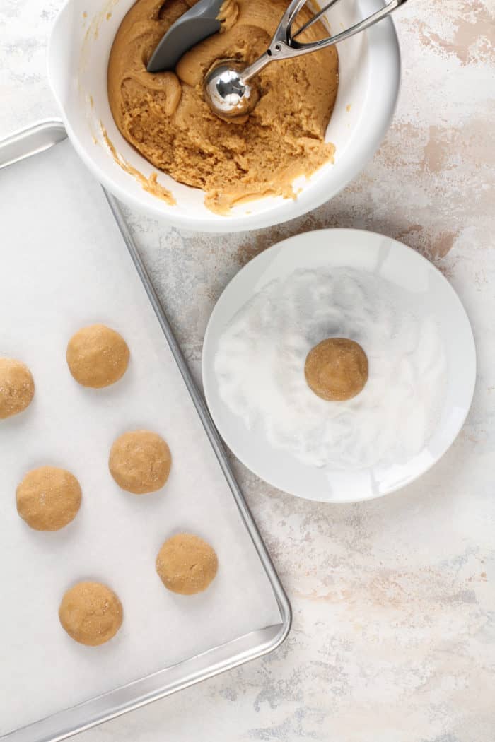 Balls of honey peanut butter cookie dough being rolled in granulated sugar and set on a baking sheet.