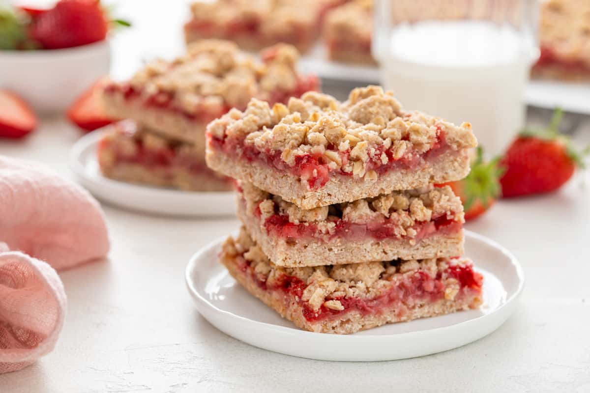 Three strawberry oatmeal bars stacked on a white plate with a glass of milk in the background.