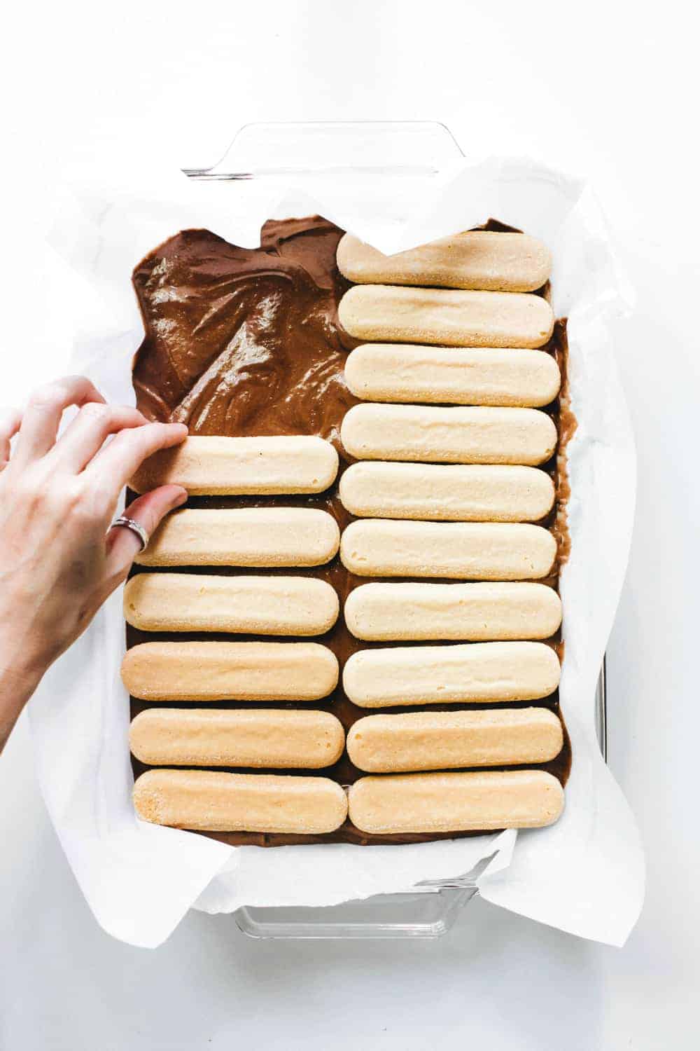 Hand placing ladyfinger cookies on top of brownie batter in a parchment-lined pan