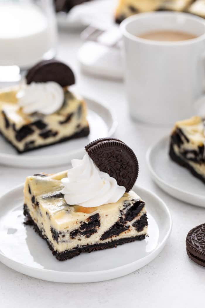 Three white plates, each holding an oreo cheesecake bar, on a white countertop with a cup of coffee in the background.