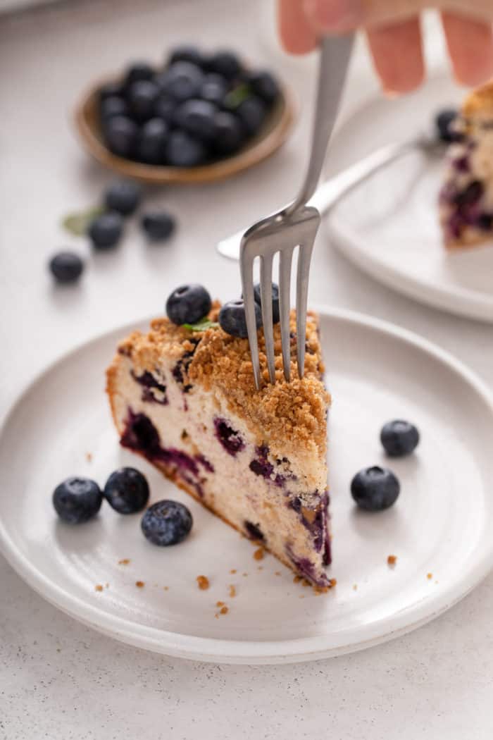 Fork about to take a bite from a slice of blueberry buckle on a white plate.