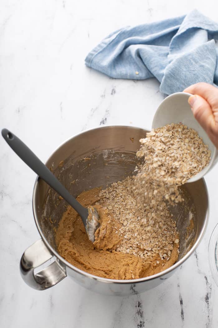 Hand pouring quick cooking oats into a mixing bowl of biscoff cookie dough