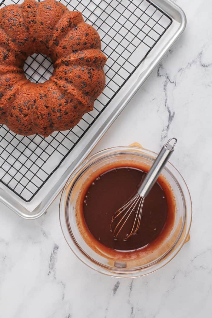 Baked chocolate chip bundt cake cooling on a wire rack next to a bowl of chocolate ganache glaze.