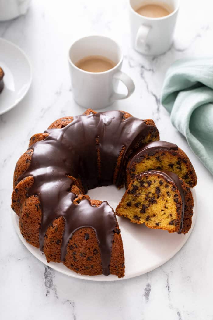 Sliced chocolate chip bundt cake set on a white cake plate. A cup of coffee is visible in the background.