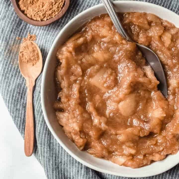 White bowl of slow cooker applesauce on a blue dishcloth next to a wooden teaspoon of cinnamon