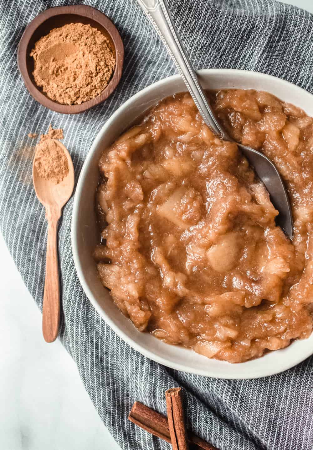 White bowl of slow cooker applesauce on a blue dishcloth next to a wooden teaspoon of cinnamon