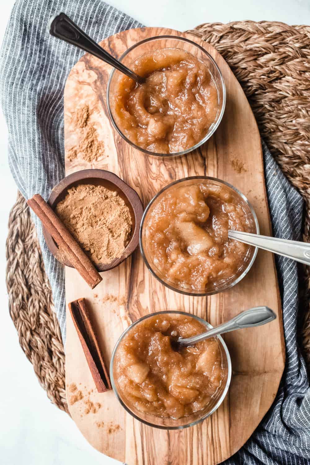 Overhead view of three bowls of crockpot applesauce on a wooden board next to cinnamon sticks