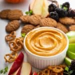 Pumpkin dip in a white bowl surrounded by fruit, pretzels, and cookies