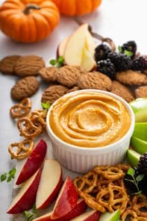 Pumpkin dip in a white bowl surrounded by fruit, pretzels, and cookies