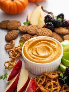 Pumpkin dip in a white bowl surrounded by fruit, pretzels, and cookies