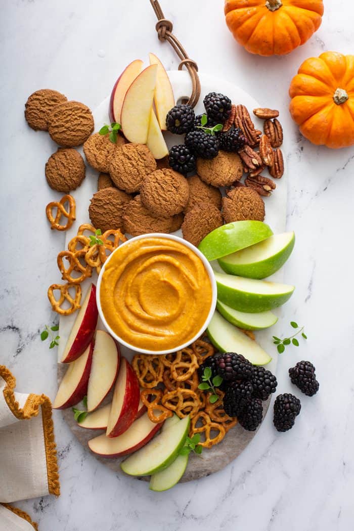 Bowl of pumpkin dip on a board surrounded by fruit, cookies, and pretzels