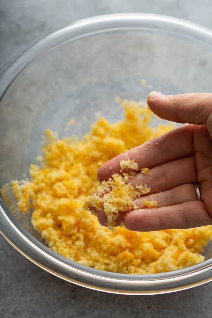 Hand showing orange zest and sugar combined together in a glass bowl