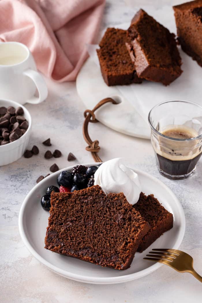 Slices of chocolate bread topped with whipped cream and berries on a white plate next to a gold fork.